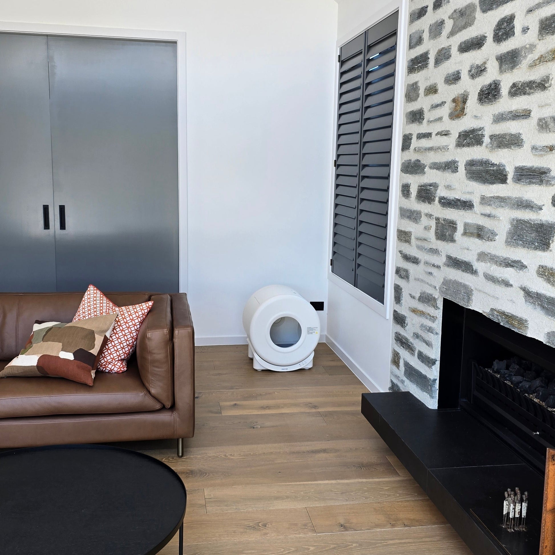 Modern living room with a cozy corner showcasing a smart automatic litter box, blending seamlessly with stylish decor, including a leather sofa, stone fireplace, and wooden flooring